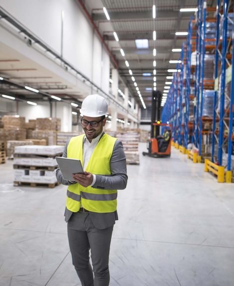 Warehouse manager walking through large storage area and holding tablet while forklift operating in background.